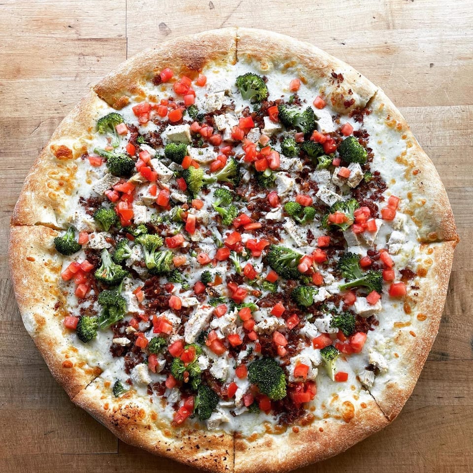 Mikey's Pizza with white sauce, chicken, broccoli, bacon bits, and diced tomatoes on a wooden table.