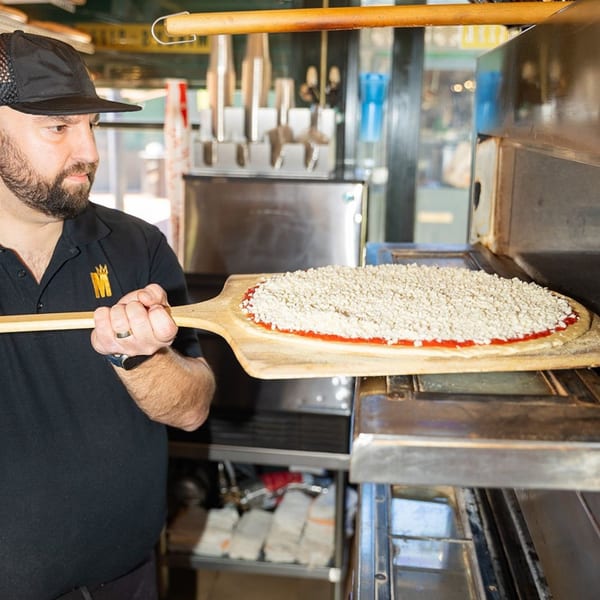 A Mikey's Pizza chef slides a raw pizza with sauce and cheese into a hot oven.