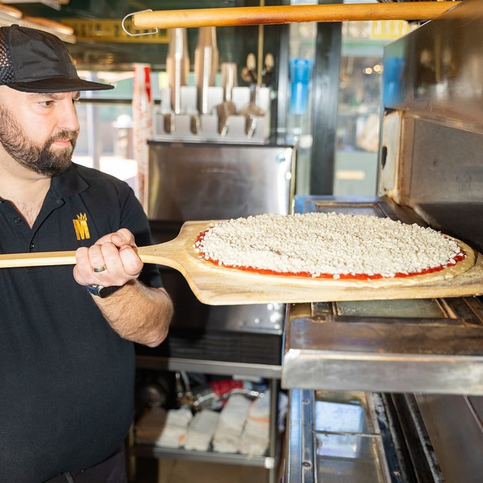 A Mikey's Pizza chef slides a raw pizza with sauce and cheese into a hot oven.