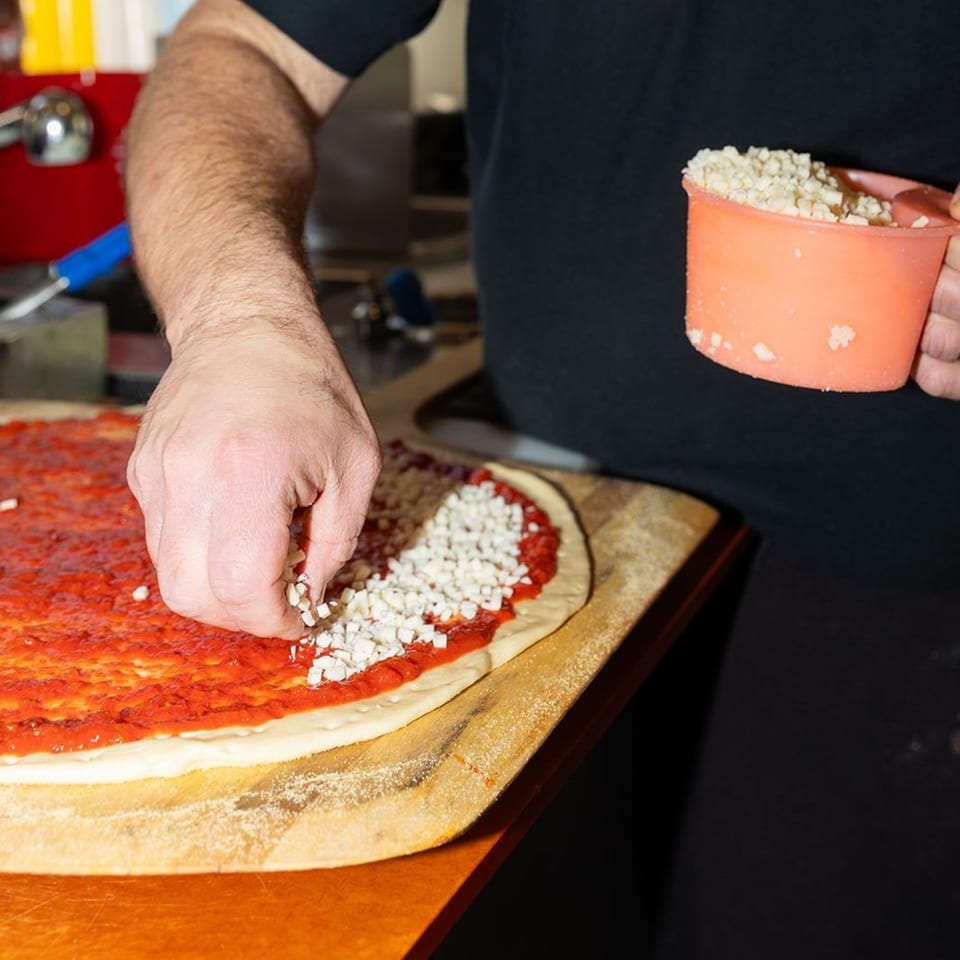 A chef's hands sprinkling cheese onto a Mikey's Pizza crust with red sauce.