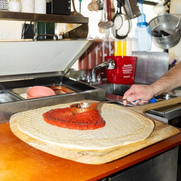 A pizza maker spreads red sauce onto a Mikey's Pizza crust on a wooden peel.